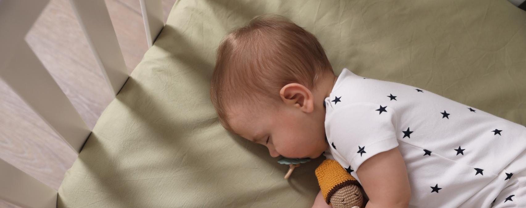 Baby sleeping in crib with toy, wearing white onesie with black stars.