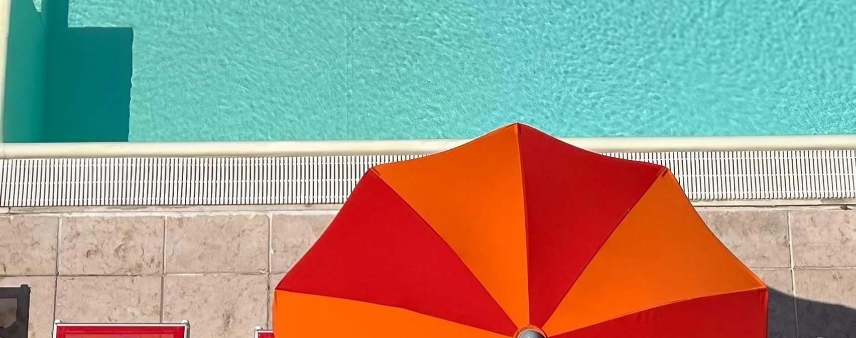 Outdoor pool with red loungers and orange umbrellas, surrounded by modern buildings.