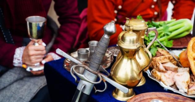 People in historical clothing with a sword and antique tableware on a table.