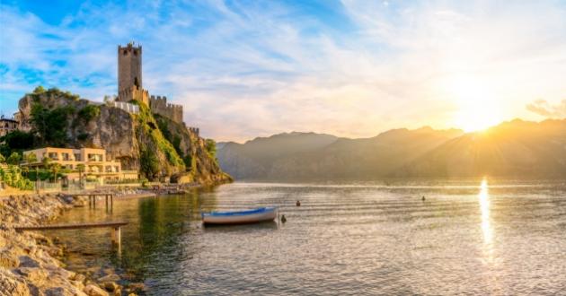 Castle by the lake at sunset with boat and mountains in the background.