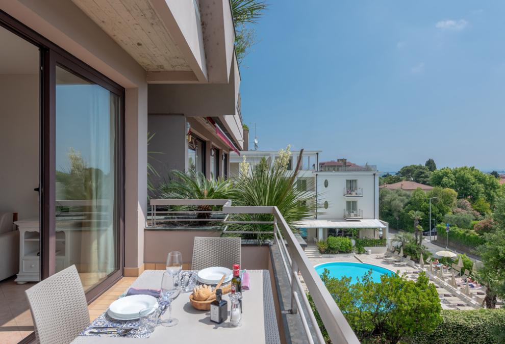 Balcone con vista piscina, tavolo apparecchiato per pranzo, giornata soleggiata.