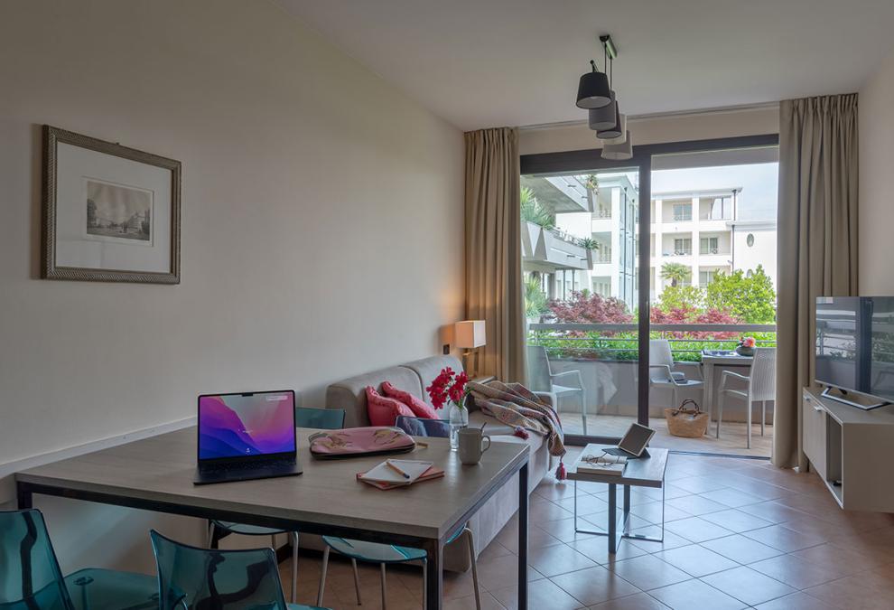 Moderno soggiorno con balcone, laptop su tavolo e vista su edifici residenziali.