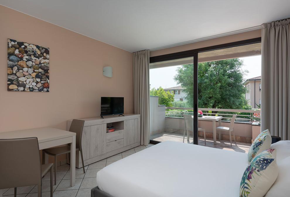 Bright room with balcony, TV, desk, and view of greenery.